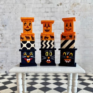 May include: Three miniature stacked boxes with Halloween themes. The top box of each stack is orange with a jack-o'-lantern face. The middle boxes are black and white with different patterns. The bottom boxes are black with a black cat face.