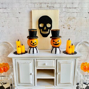 K&ouml;nnte beinhalten: Zwei Miniatur-Jack-o'-Lantern-Kerzenhalter mit schwarzen Zylindern, eine schwarze Sch&auml;delsilhouette auf einer Holztafel und gelbe Kerzen auf einem wei&szlig;en, abgenutzten Schrank mit einem schwarz-wei&szlig; karierten Boden.