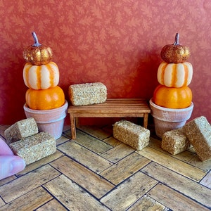 Op de afbeelding: Twee miniatuurstapels pompoenen, één oranje en één met een gouden glittertop, staan op een houten bankje met hooibalen op een houten vloer.