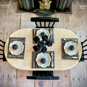 May include: A black and white table setting with three place settings, each with a white plate, black and gold silverware, and a black pumpkin-shaped centerpiece. The table is set on a wooden floor with a fireplace in the background.