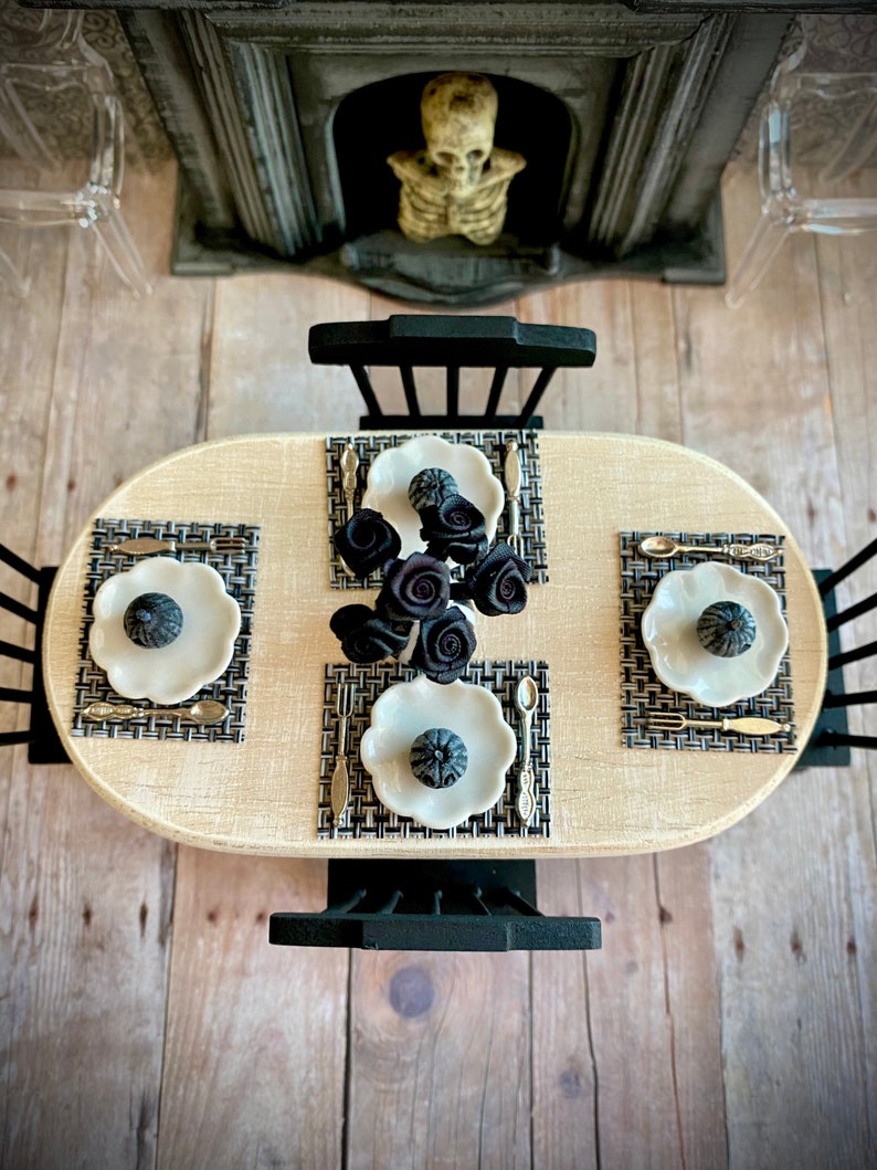 May include: A black and white table setting with three place settings, each with a white plate, black and gold silverware, and a black pumpkin-shaped centerpiece. The table is set on a wooden floor with a fireplace in the background.