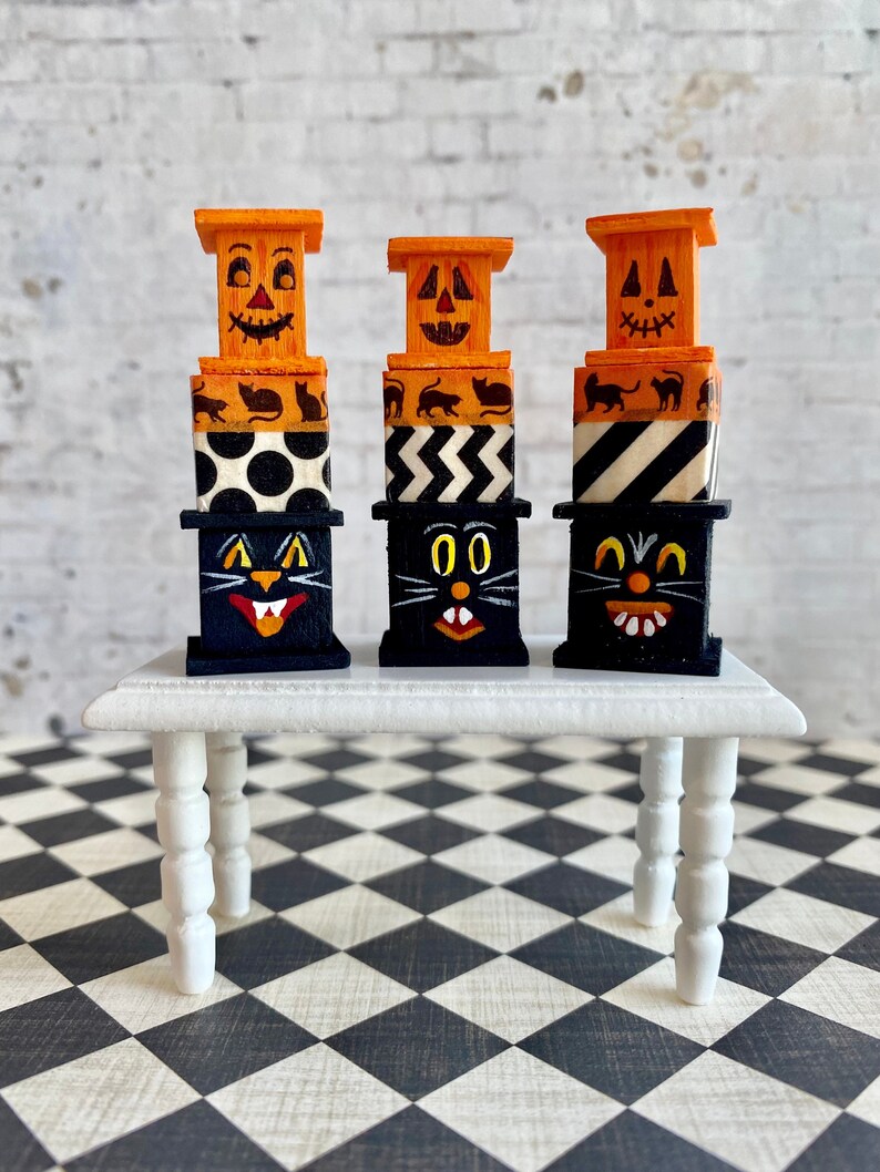 May include: Three miniature stacked boxes with Halloween themes. The top box of each stack is orange with a jack-o'-lantern face. The middle boxes are black and white with different patterns. The bottom boxes are black with a black cat face.