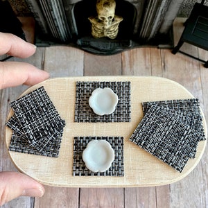 May include: A set of four black and white woven placemats with white ceramic plates on top. The placemats are on a wooden table.
