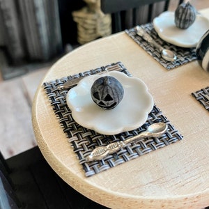 May include: A black and white checkered placemat with a white plate and a black pumpkin-shaped candle on top. A silver spoon is on the placemat.