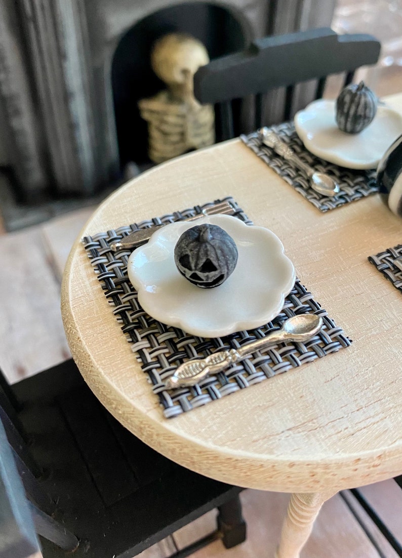 May include: A black and white checkered placemat with a white plate and a black pumpkin-shaped candle on top. A silver spoon is on the placemat.