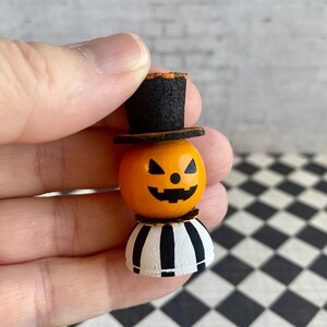 K&ouml;nnte beinhalten: Eine kleine Holz-K&uuml;rbisfigur mit einem schwarzen Zylinder und einem schwarz-wei&szlig; gestreiften Sockel. Der K&uuml;rbis hat ein geschnitztes Jack-o'-Lantern-Gesicht.
