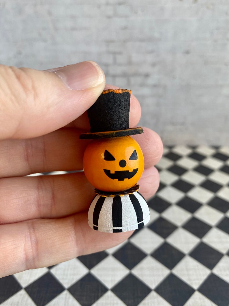 K&ouml;nnte beinhalten: Eine kleine Holz-K&uuml;rbisfigur mit einem schwarzen Zylinder und einem schwarz-wei&szlig; gestreiften Sockel. Der K&uuml;rbis hat ein geschnitztes Jack-o'-Lantern-Gesicht.