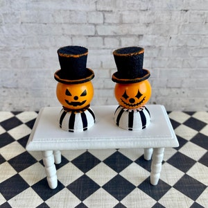 K&ouml;nnte beinhalten: Zwei Miniatur-Jack-o'-Lantern-Figuren mit schwarzen Zylindern und gestreiften Sockeln, die auf einem wei&szlig;en Tisch mit einem schwarz-wei&szlig; karierten Boden stehen.