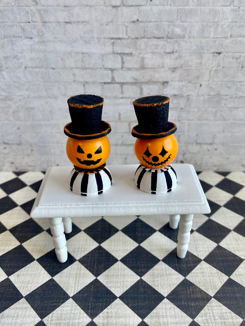 K&ouml;nnte beinhalten: Zwei Miniatur-Jack-o'-Lantern-Figuren mit schwarzen Zylindern und gestreiften Sockeln, die auf einem wei&szlig;en Tisch mit einem schwarz-wei&szlig; karierten Boden stehen.
