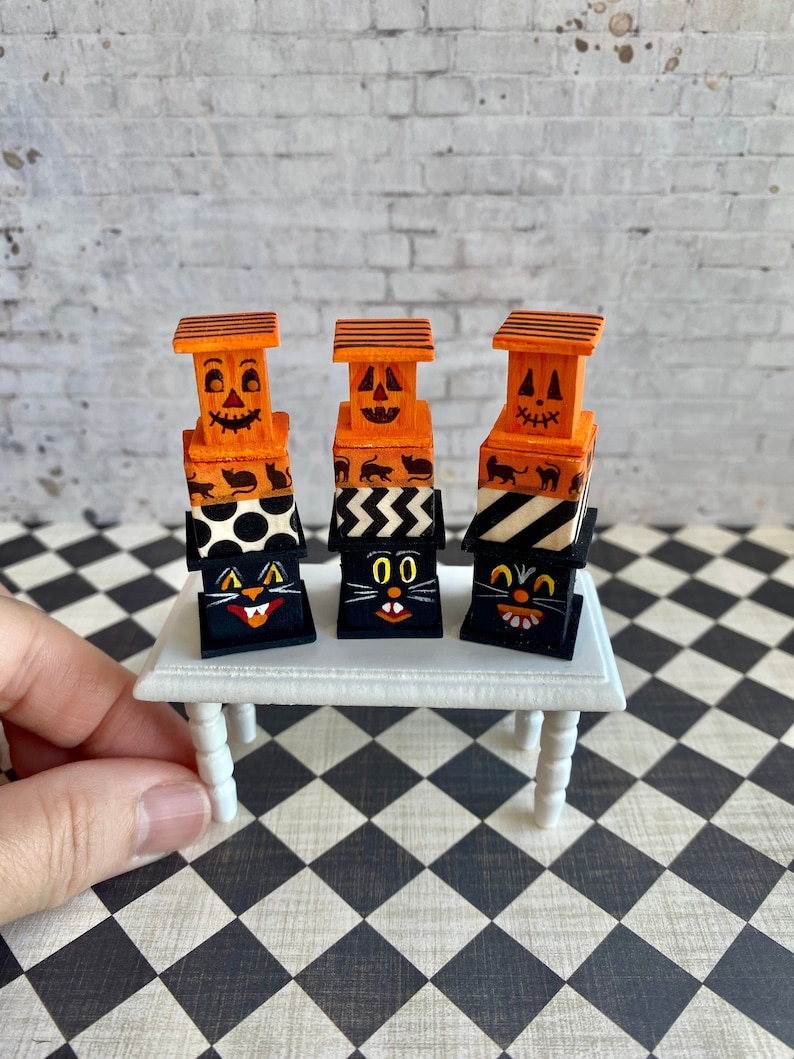 May include: Three stacked wooden boxes with Halloween-themed designs. The top box features a jack-o'-lantern face, the middle box has a black cat face, and the bottom box has a black cat face with a white stripe. The boxes are painted in orange, black, and white.