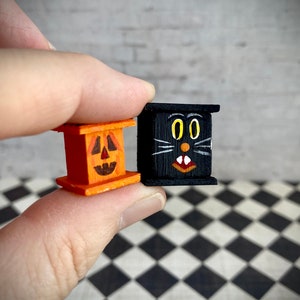 May include: Two small wooden Halloween decorations. One is a pumpkin with a carved face and the other is a black cat with yellow eyes and a white mouth.