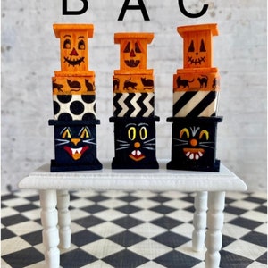 May include: Three stacked Halloween-themed decorations, each featuring a black cat face, orange jack-o'-lantern face, and black and white patterned boxes. The decorations are arranged on a white table with black and white checkered floor.
