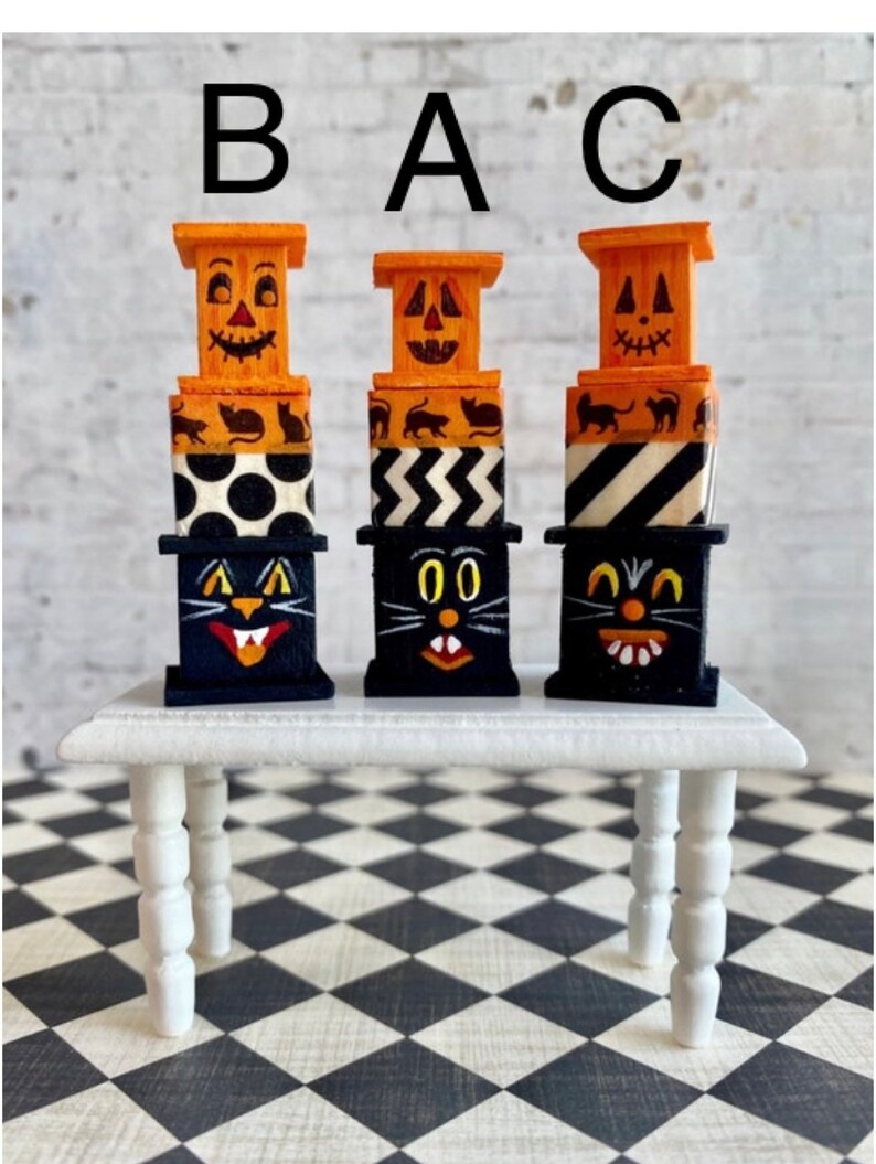 May include: Three stacked Halloween-themed decorations, each featuring a black cat face, orange jack-o'-lantern face, and black and white patterned boxes. The decorations are arranged on a white table with black and white checkered floor.