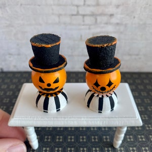 K&ouml;nnte beinhalten: Zwei Miniatur-Jack-o'-Lantern-Figuren mit schwarzen Zylindern und gestreiften Sockeln auf einem wei&szlig;en Tisch. Die Jack-o'-Lanterns haben orange Gesichter mit schwarzen Augen, Nasen und M&uuml;ndern.