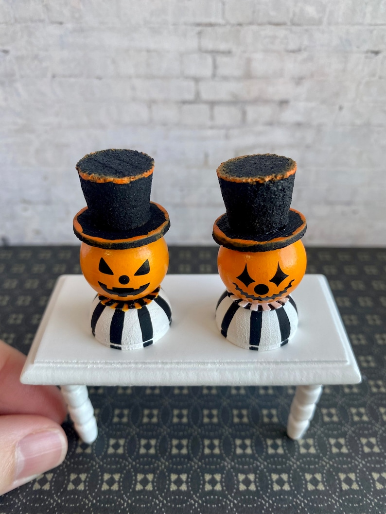 K&ouml;nnte beinhalten: Zwei Miniatur-Jack-o'-Lantern-Figuren mit schwarzen Zylindern und gestreiften Sockeln auf einem wei&szlig;en Tisch. Die Jack-o'-Lanterns haben orange Gesichter mit schwarzen Augen, Nasen und M&uuml;ndern.