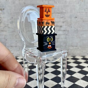 May include: A miniature clear plastic chair with a stack of four wooden boxes on the seat. The boxes are decorated with Halloween themes, including a jack-o'-lantern face, black cats, and a black cat with a white face.