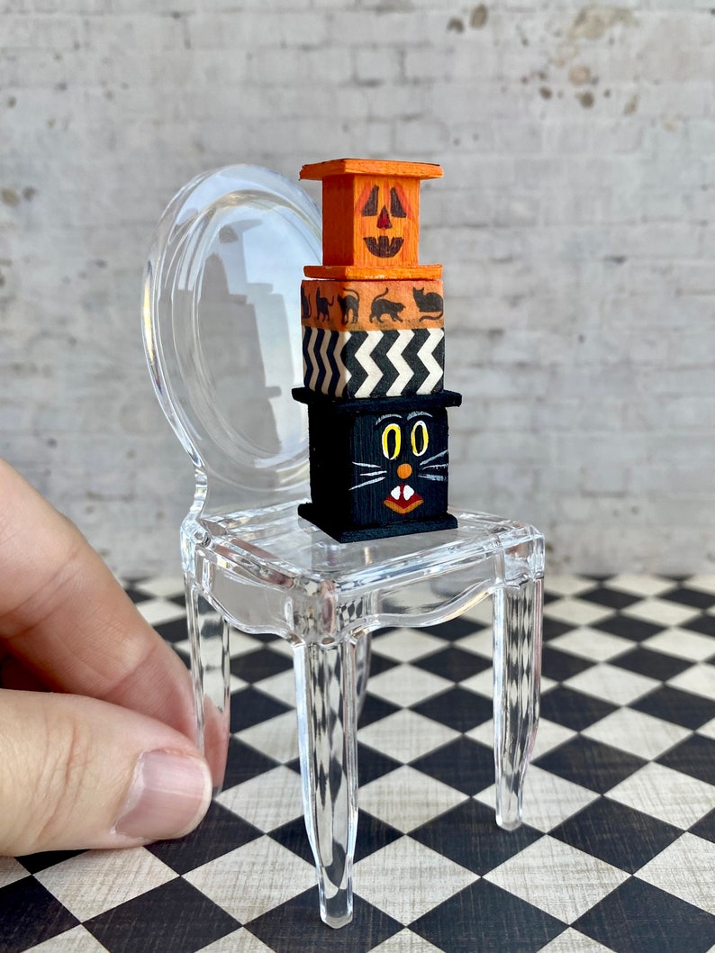 May include: A miniature clear plastic chair with a stack of four wooden boxes on the seat. The boxes are decorated with Halloween themes, including a jack-o'-lantern face, black cats, and a black cat with a white face.