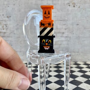 May include: A miniature Halloween-themed stack of wooden boxes with a black cat face, black and white stripes, and a jack-o'-lantern face. The stack is sitting on a clear plastic chair.