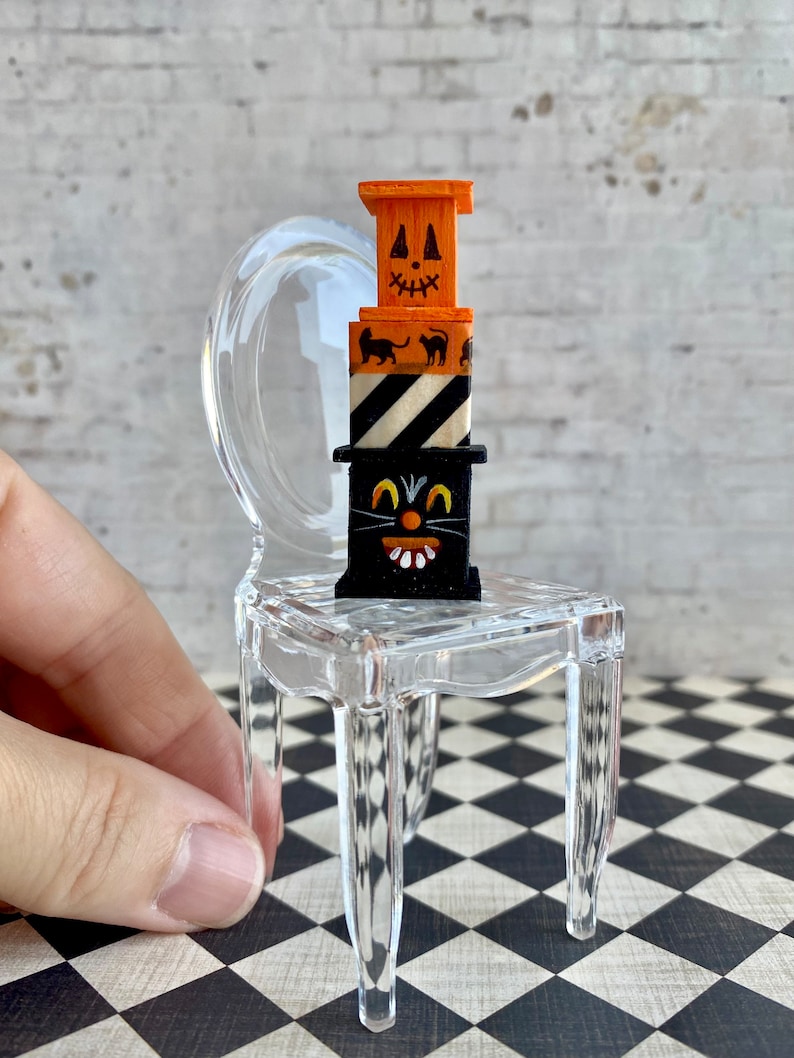 May include: A miniature Halloween-themed stack of wooden boxes with a black cat face, black and white stripes, and a jack-o'-lantern face. The stack is sitting on a clear plastic chair.