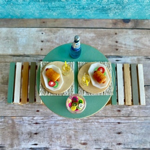 May include: A miniature picnic scene with a green table, two white plates with hot dogs and salad, two glasses of lemonade, a bottle of blue soda, and a bowl of fruit salad.