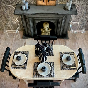 May include: A miniature dining table setting with black and white placemats, plates, and silverware. A vase with black roses sits in the center of the table. The table is surrounded by black chairs and a fireplace with a skeleton in the background.