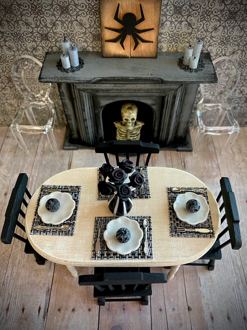 May include: A miniature dining table setting with black and white placemats, plates, and silverware. A vase with black roses sits in the center of the table. The table is surrounded by black chairs and a fireplace with a skeleton in the background.