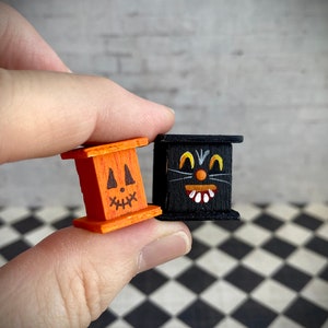 May include: Two small wooden boxes with painted faces. One box is orange with a jack-o'-lantern face and the other is black with a black cat face.