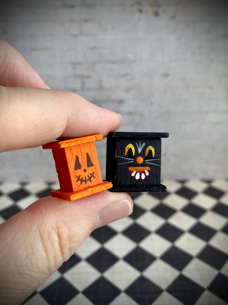May include: Two small wooden boxes with painted faces. One box is orange with a jack-o'-lantern face and the other is black with a black cat face.