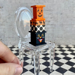 May include: A stack of three miniature wooden boxes decorated with Halloween themes. The top box is orange and features a jack-o'-lantern face. The middle box is black and white polka dot. The bottom box is black and features a black cat face. The boxes are sitting on a clear plastic chair.