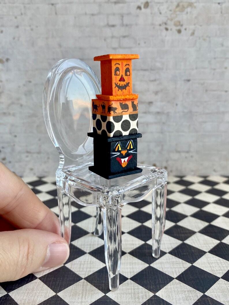 May include: A stack of three miniature wooden boxes decorated with Halloween themes. The top box is orange and features a jack-o'-lantern face. The middle box is black and white polka dot. The bottom box is black and features a black cat face. The boxes are sitting on a clear plastic chair.