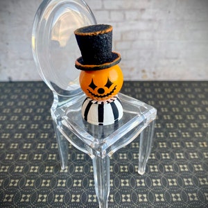 K&ouml;nnte beinhalten: Ein kleiner, orangefarbener K&uuml;rbis mit einem schwarzen Zylinder und einem schwarz-wei&szlig; gestreiften K&ouml;rper sitzt auf einem durchsichtigen Plastikstuhl. Der Stuhl steht auf einer schwarz-wei&szlig; gemusterten Oberfl&auml;che.
