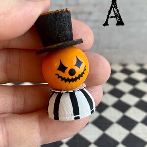 K&ouml;nnte beinhalten: Eine kleine, orangefarbene Figur in Form eines K&uuml;rbisses mit einem schwarzen Zylinder, einem schwarz-wei&szlig; gestreiften Rock und einem bemalten K&uuml;rbisgesicht. Die Figur wird in einer Hand gehalten.