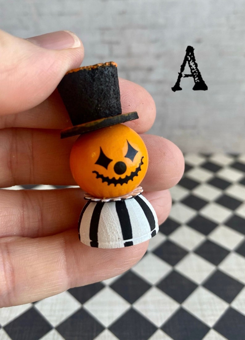 K&ouml;nnte beinhalten: Eine kleine, orangefarbene Figur in Form eines K&uuml;rbisses mit einem schwarzen Zylinder, einem schwarz-wei&szlig; gestreiften Rock und einem bemalten K&uuml;rbisgesicht. Die Figur wird in einer Hand gehalten.