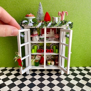 May include: A white cabinet with glass doors decorated for Christmas. The cabinet is filled with miniature Christmas decorations, including a snowman, a Christmas tree, and a Santa Claus figurine. The cabinet is sitting on a black and white checkered floor.