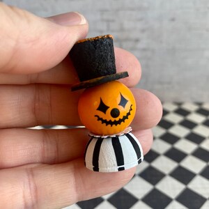 K&ouml;nnte beinhalten: Ein kleiner orangefarbener K&uuml;rbis mit einem schwarzen K&uuml;rbisgesicht, der einen schwarzen Zylinder mit einem wei&szlig;en Band und einen schwarz-wei&szlig; gestreiften Rock tr&auml;gt.