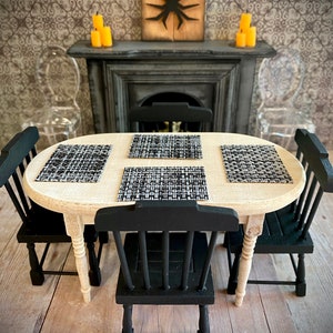 May include: A miniature dining table set with four black chairs and a light wood oval table. The table is set with black and white placemats. The background features a fireplace with a black silhouette of a spider on a wooden plaque.
