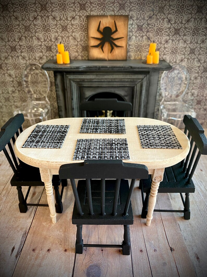 May include: A miniature dining table set with four black chairs and a light wood oval table. The table is set with black and white placemats. The background features a fireplace with a black silhouette of a spider on a wooden plaque.
