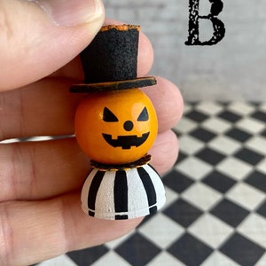 K&ouml;nnte beinhalten: Eine kleine, h&ouml;lzerne Jack-o'-Lantern-Figur mit einem schwarzen Zylinder, einem orangefarbenen Gesicht und einem schwarz-wei&szlig; gestreiften Sockel. Die Figur wird in einer Hand gehalten.