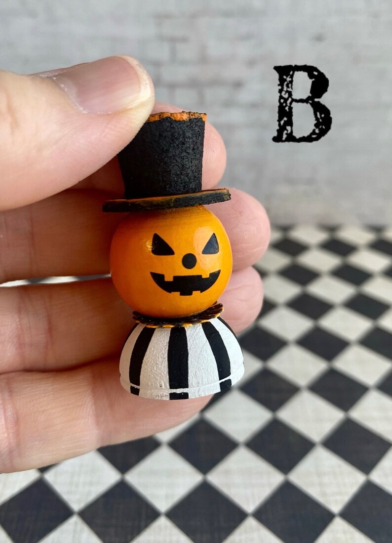 K&ouml;nnte beinhalten: Eine kleine, h&ouml;lzerne Jack-o'-Lantern-Figur mit einem schwarzen Zylinder, einem orangefarbenen Gesicht und einem schwarz-wei&szlig; gestreiften Sockel. Die Figur wird in einer Hand gehalten.