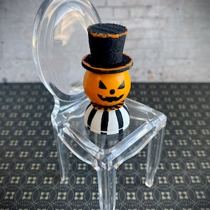 K&ouml;nnte beinhalten: Ein kleiner, orangefarbener K&uuml;rbis mit einem schwarzen Zylinder sitzt auf einem durchsichtigen Plastikstuhl. Der K&uuml;rbis hat ein geschnitztes Jack-o'-Lantern-Gesicht und einen schwarz-wei&szlig; gestreiften Sockel.