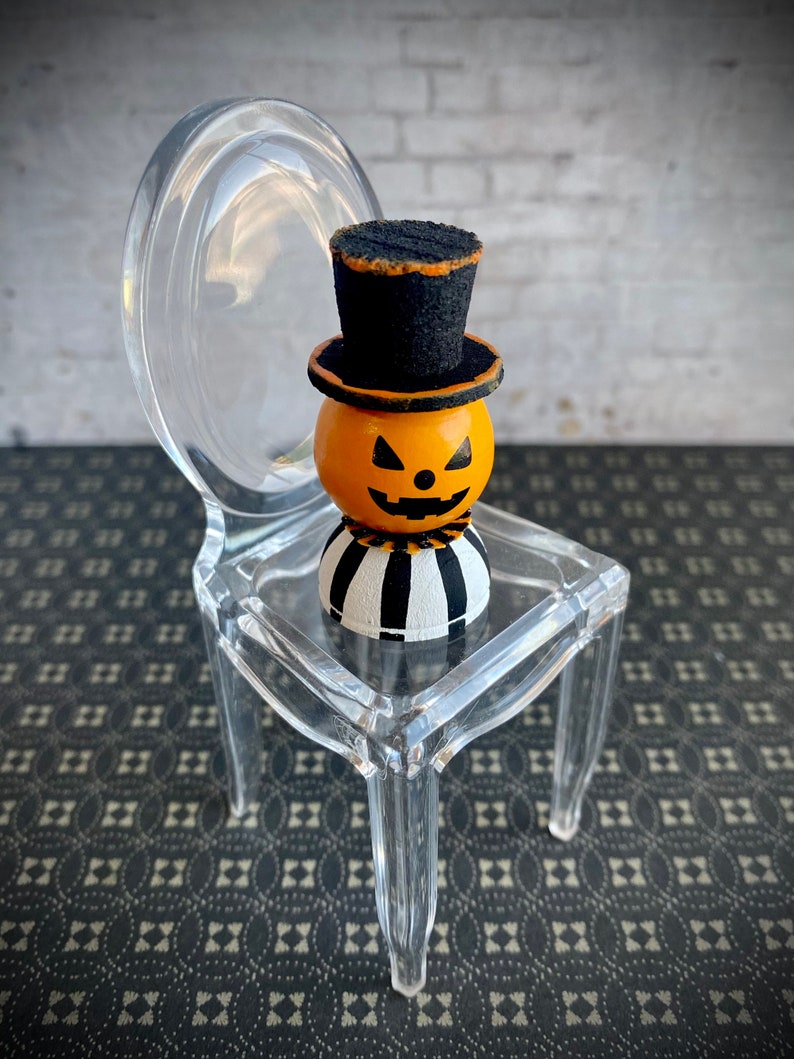 K&ouml;nnte beinhalten: Ein kleiner, orangefarbener K&uuml;rbis mit einem schwarzen Zylinder sitzt auf einem durchsichtigen Plastikstuhl. Der K&uuml;rbis hat ein geschnitztes Jack-o'-Lantern-Gesicht und einen schwarz-wei&szlig; gestreiften Sockel.