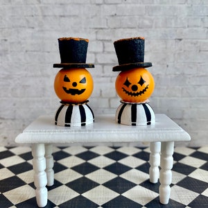 K&ouml;nnte beinhalten: Zwei Miniatur-Jack-o'-Lantern-Figuren mit schwarzen Zylindern und gestreiften Sockeln, die auf einem wei&szlig;en Tisch mit gedrehten Beinen stehen. Der Tisch steht auf einem schwarz-wei&szlig; karierten Boden.