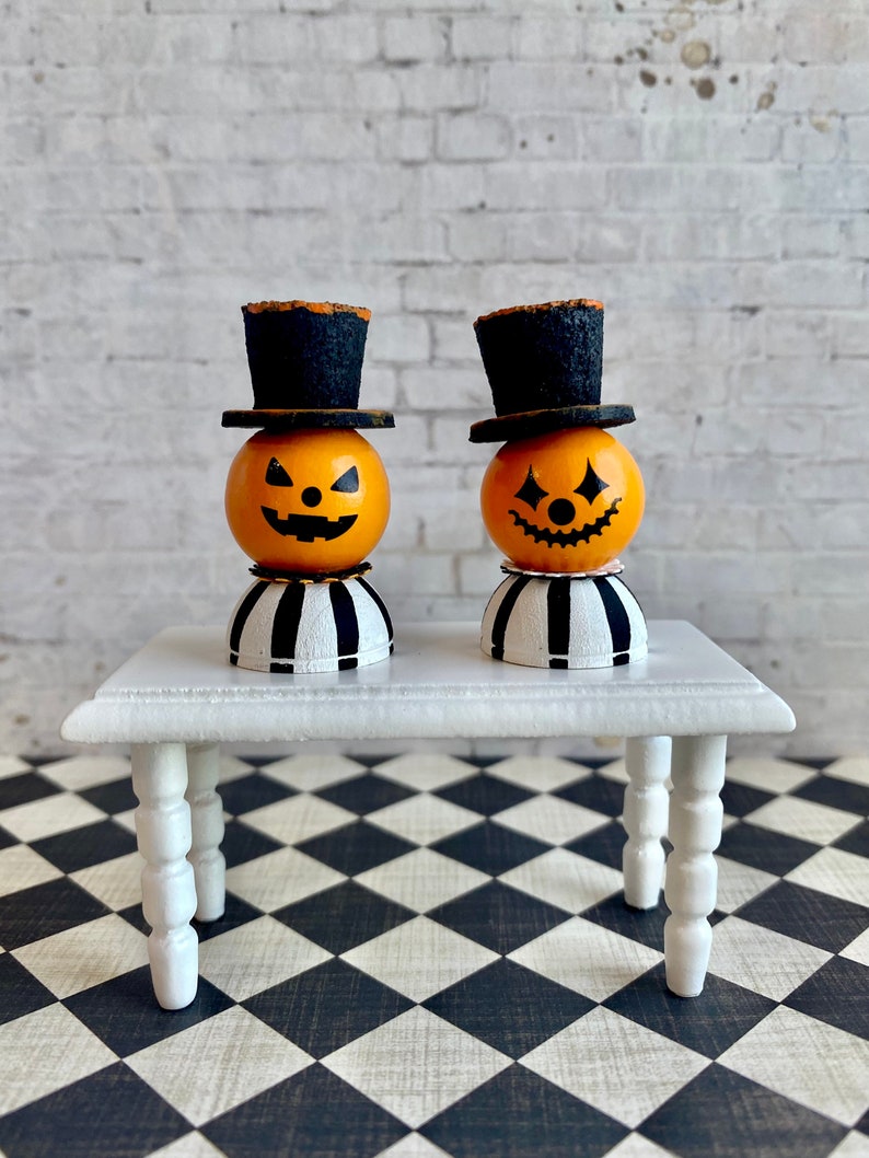 K&ouml;nnte beinhalten: Zwei Miniatur-Jack-o'-Lantern-Figuren mit schwarzen Zylindern und gestreiften Sockeln, die auf einem wei&szlig;en Tisch mit gedrehten Beinen stehen. Der Tisch steht auf einem schwarz-wei&szlig; karierten Boden.
