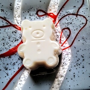 Puede incluir: Un jabón blanco con forma de oso se encuentra sobre una superficie azul claro con motas negras y detalles rojos. El jabón tiene un acabado liso y brillante con detalles de botones y ojos. El fondo tiene una apariencia blanca texturizada.