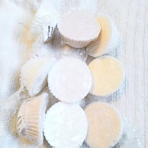 May include: Several round soaps, wrapped in clear plastic, in shades of white and cream, are arranged on a white towel. Some are in paper cups. The text "Milk Soap Breeze" is visible on one wrapper.