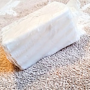 May include: A rectangular block of white soap wrapped in clear plastic. The soap is sitting on a textured, beige towel. The soap appears to be unscented.