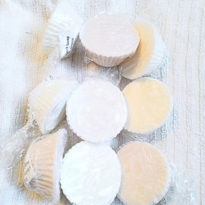 Peut inclure: Plusieurs savons ronds, emballés dans du plastique transparent, dans des tons de blanc et de crème. Certains sont dans des coupelles en papier. Le texte "Milk Soap Breeze" est visible sur un emballage. Les savons sont disposés sur une surface blanche texturée.