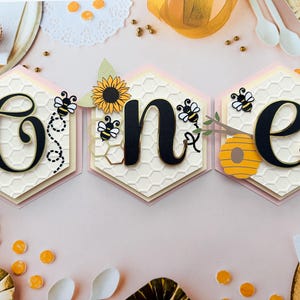 May include: A decorative banner spelling "ONE" in black script, with a bee and sunflower theme. The banner is made of hexagonal shapes with a honeycomb pattern, and is decorated with bees, sunflowers, and a beehive.