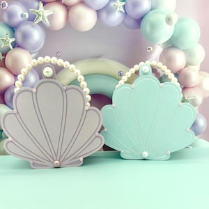 May include: Two shell-shaped purses, one in light lavender and one in mint green, each featuring a pearl-beaded handle and a single pearl accent at the base. The purses have a textured surface with embossed lines radiating from the bottom center.