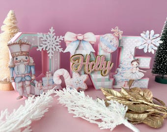 May include: A pink and white Christmas-themed decoration spelling out the name "Holly". The letters are decorated with a nutcracker, snowflakes, a bow, and a ballerina. Additional decorations include a small Christmas tree and gold leaves.